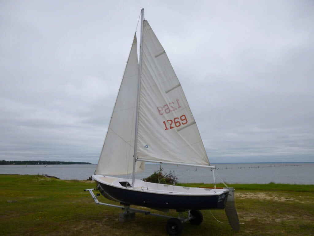 Photo of our first sail boat, 13 foot Chrysler, sitting on a dolly in the grass in front of the Mobile Bay on a cloudy day with both sails up.