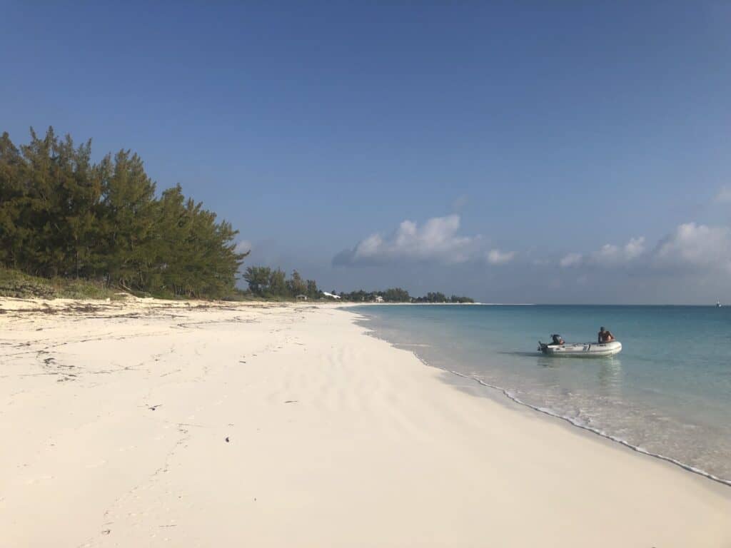 View of the beach, with trees along the upper edge of the beach and a dinghy anchored in the water on a sunny morning.