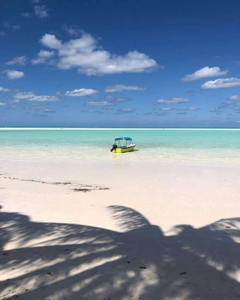Clear blue, turquoise, flat water with a sandbar out in the distance. There is a small yellow boat with green canvas anchored in the shallow water, and the shadow of a palm tree in the foreground.