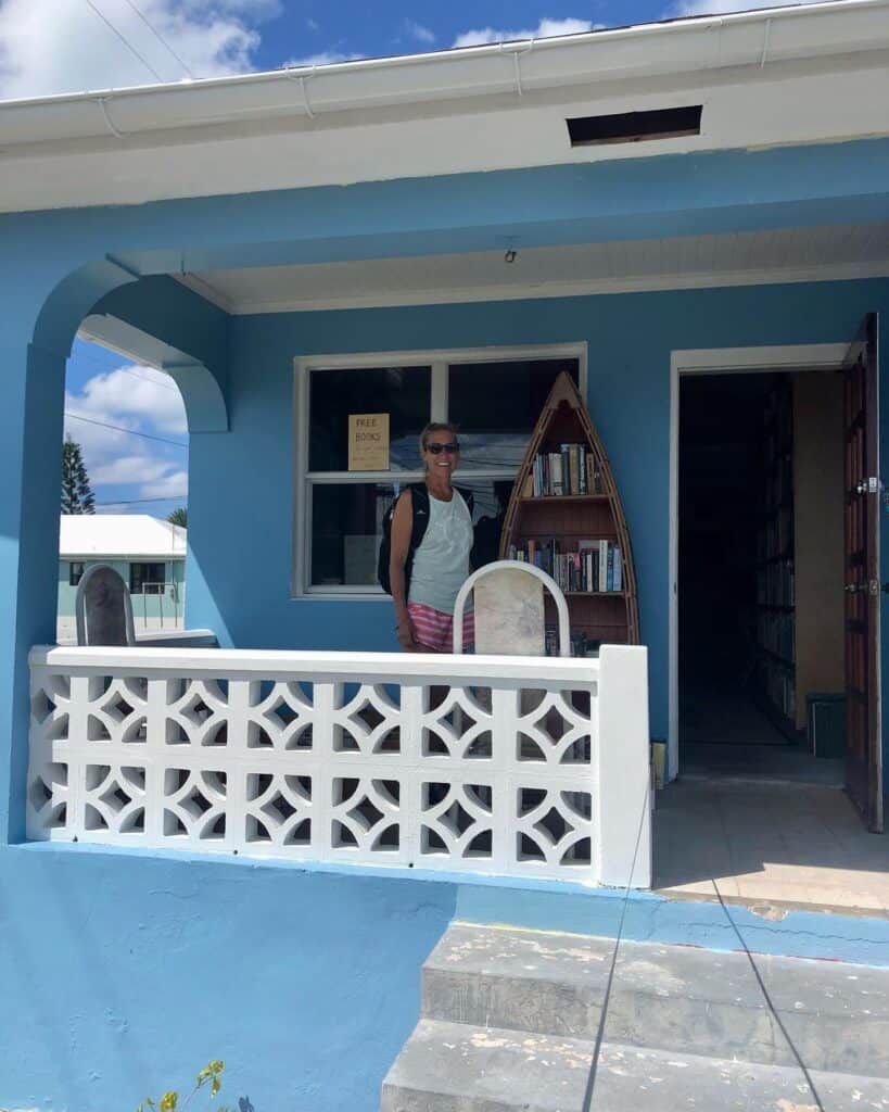 I am standing on the porch of a blue house with white accents that is the bookstore/library in Spanish Wells. There is a book shelf on the porch shaped like a boat with books on it. The door is open and the room inside is full of more books.