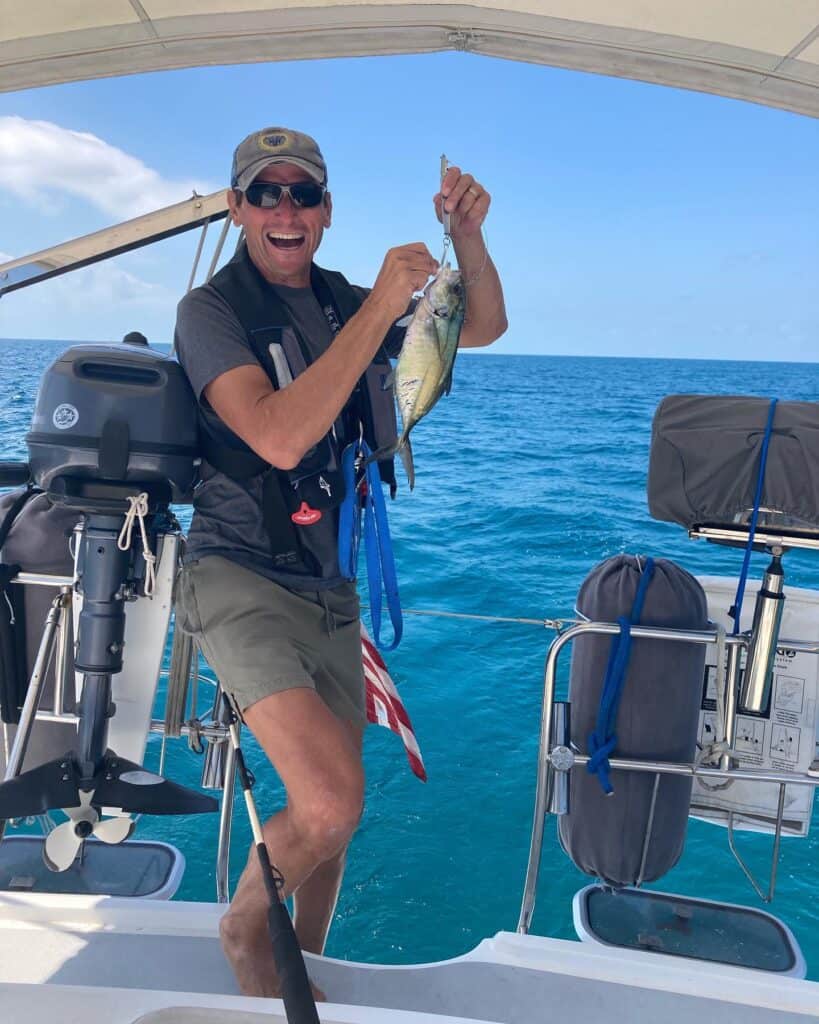 Scott on the stern of the sailboat holding the blue runner fish he just caught while we were under sail on the Bahamas Banks. Scott is wearing his life jacket and has a huge smile on his face.