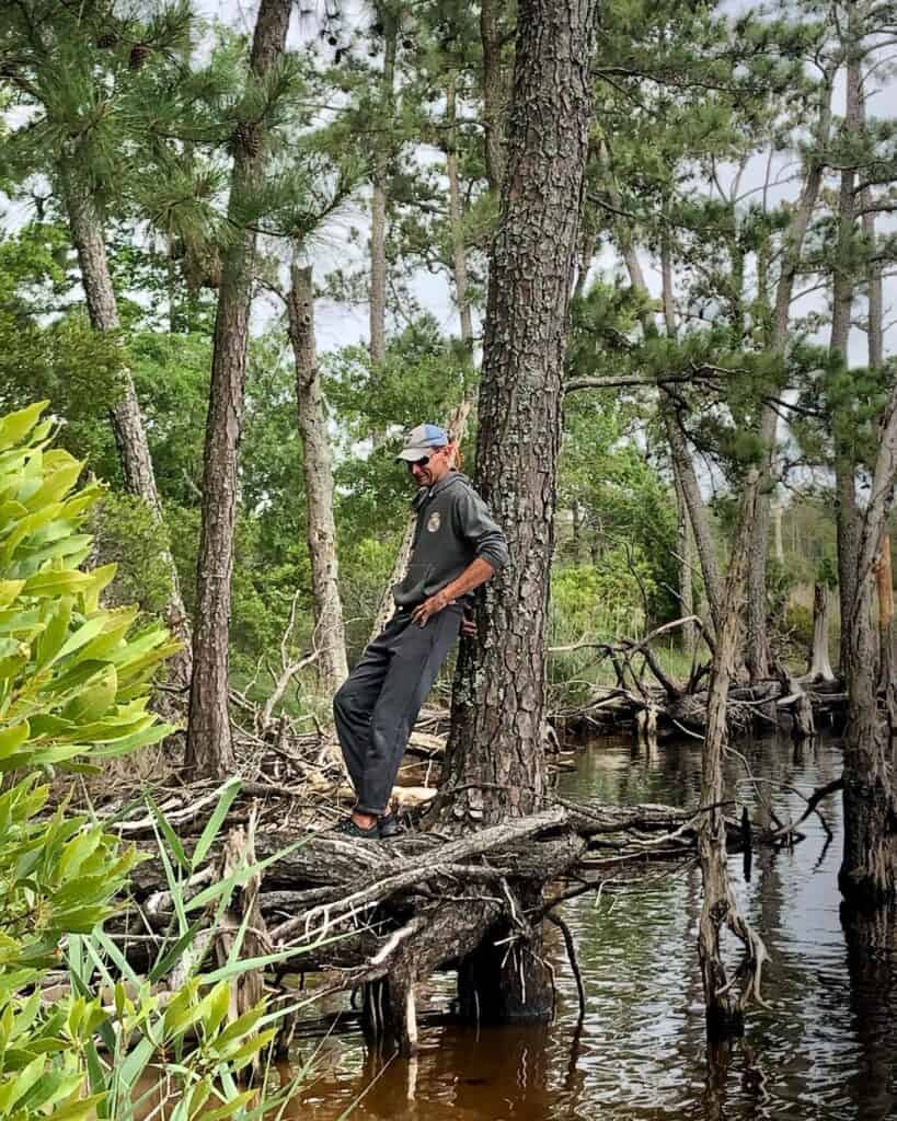 Scott in a gray sweatshirt and black sweatpants smiling and leaning against a tree over the water. He is watching our dog Mangaux explore this tiny beach on his walk.
