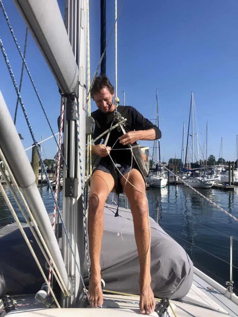 We save money sailing on a budget by inspecting our own mast and rigging. Scott is in the bosun chair at the bottom of the mast getting ready to be hoisted up to inspect the mast and rigging on a sunny day.