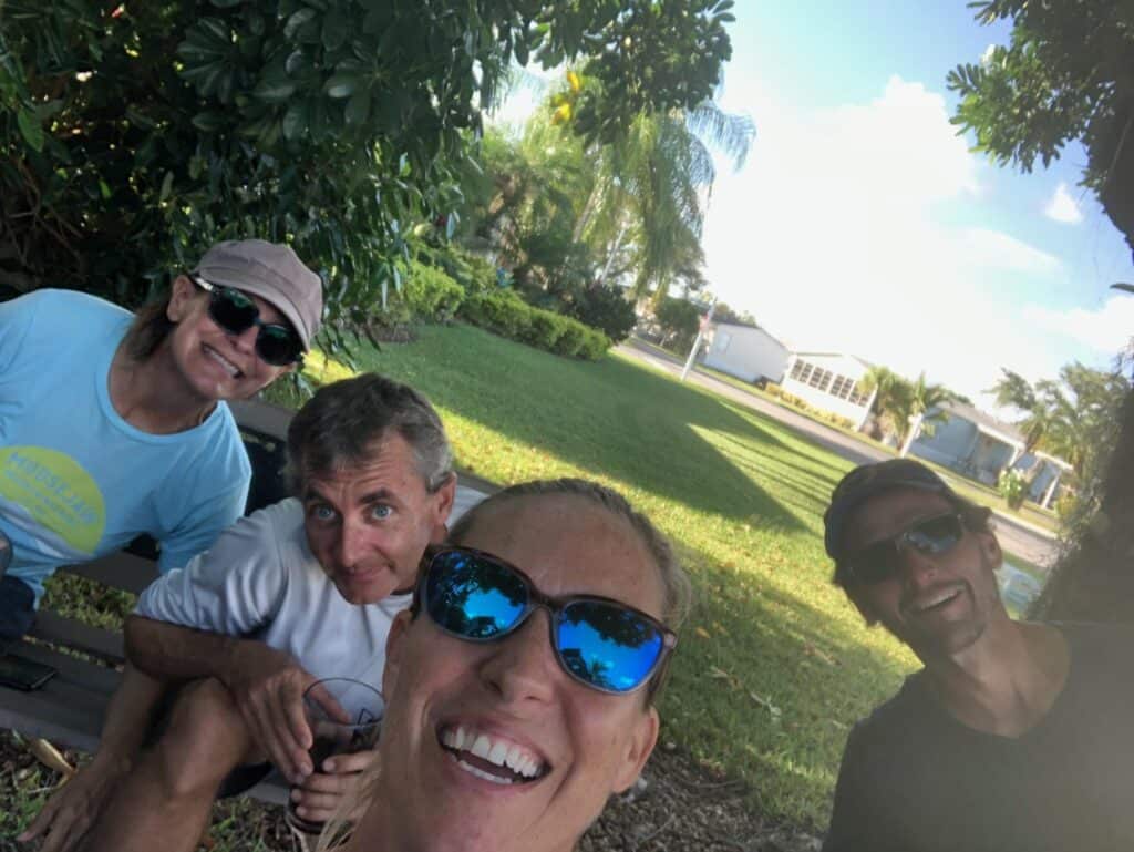 Casie, Tyler, me (selfie) and Scott sitting outside in the yard at Scott's dad's house on a sunny day for this sailing interview. Grass, houses and palm trees are in the background.