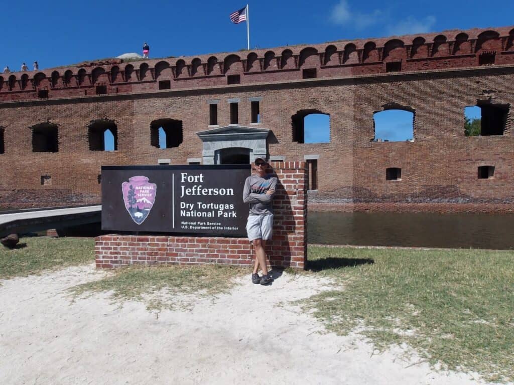 Tyler is standing in front of the Fort Jefferson - Dry Tortugas National Park sign at Fort Jefferson on a sunny day. He is wearing shorts and a gray long sleeved shirt.