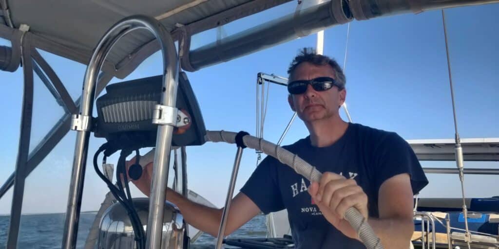 Tyler sailing SV Vagari on a clear sunny day. Tyler is not smiling (he never does for the camera!) and wearing a navy blue t shirt.