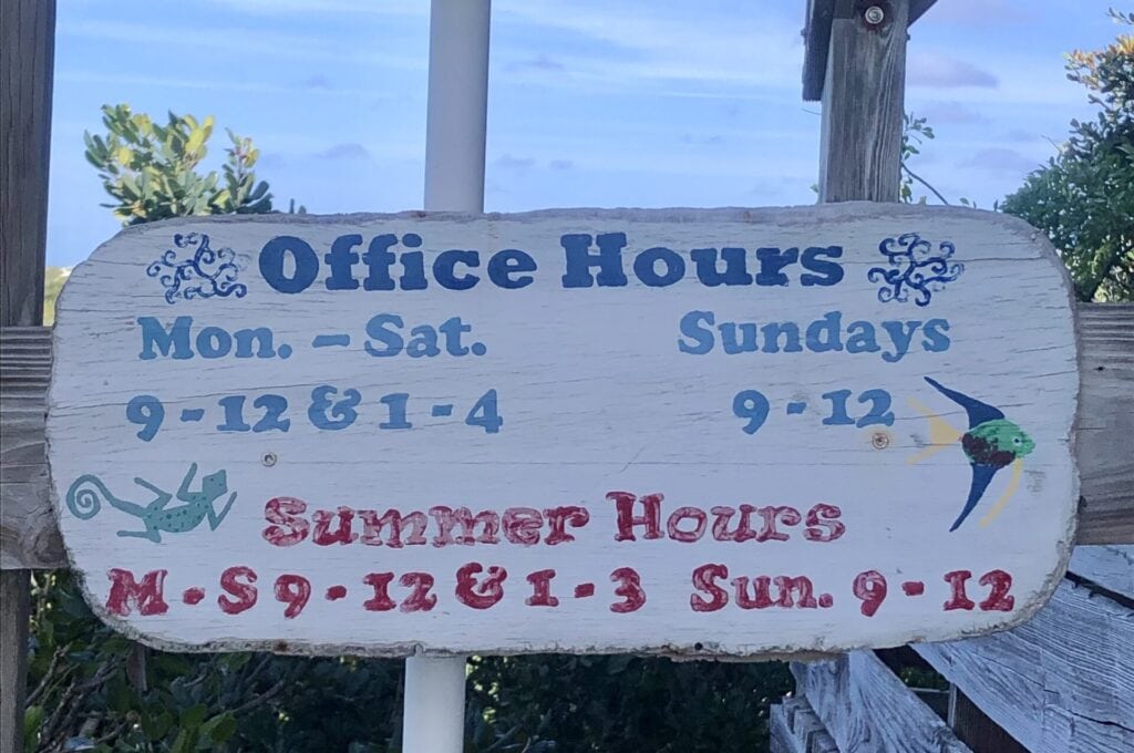 A colorful wood sign displays the office hours at the park office in Warderick Wells.