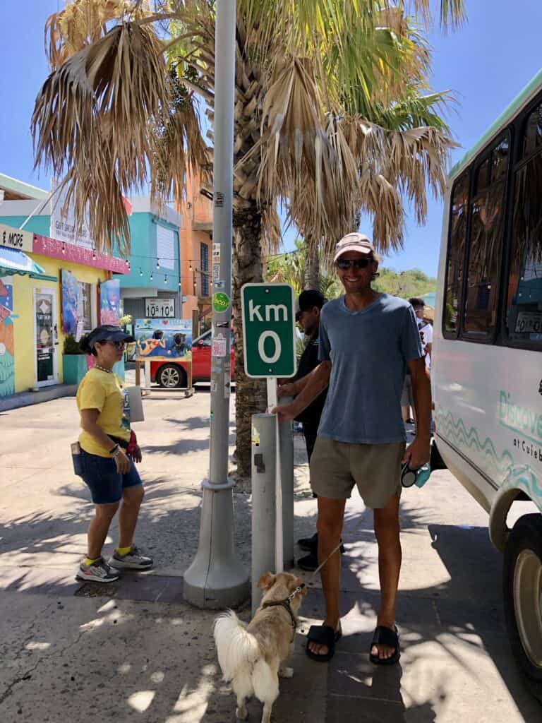 Scott at KM marker 0 on the waterfront street in Culebra next to palm trees and colorful buildings.