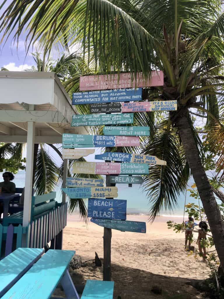 Lower Bay Beach at Keegan's. A post with colorful signs near palm trees are in front of the beach at the restaurant.