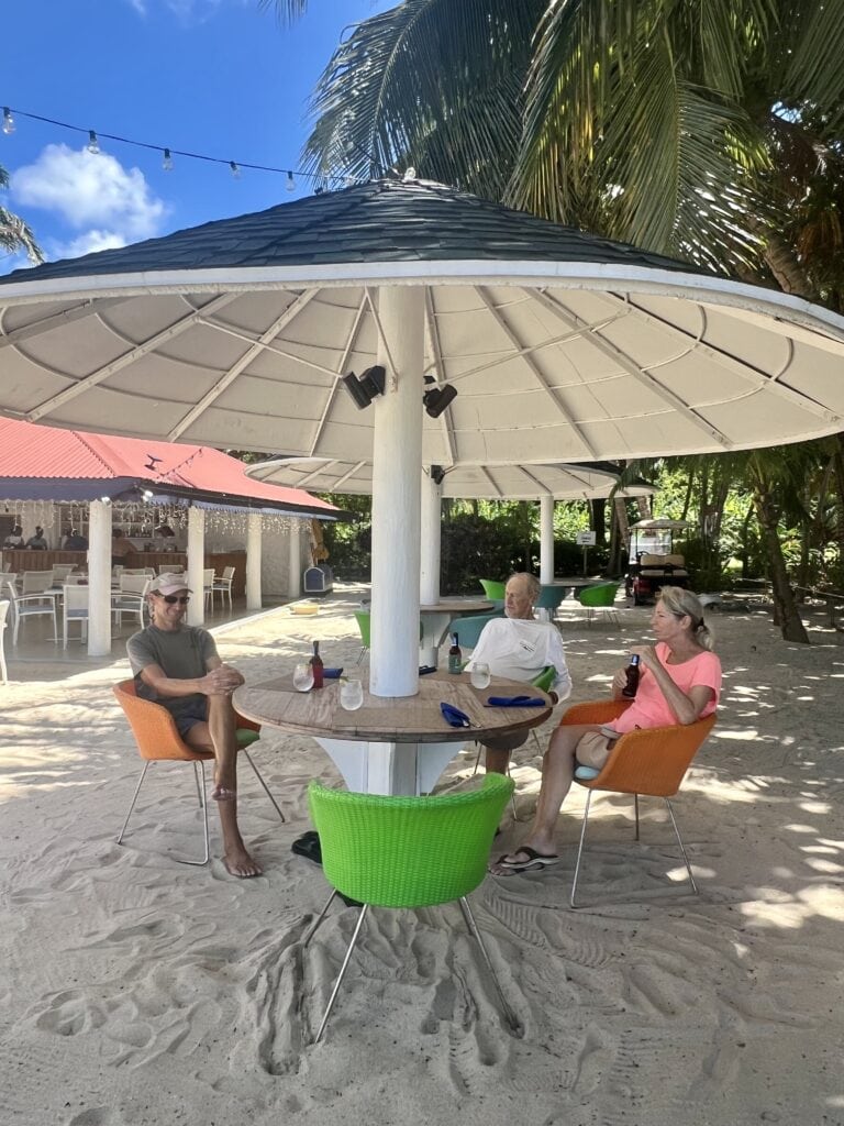 Scott, Scott's Dad, and Winnie are sitting at a table with a thatched umbrella and colorful orange and green chairs in the sand at Savvy's Restaurant. Savvy's is one of the best restaurants in Grenada for its watermelon tomato salad.