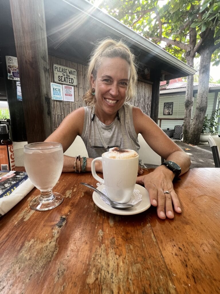 I am sitting outside at Victory Bar with a coffee and water in front of me for my birthday breakfast. Behind me is the bamboo wall of the restaurant and a big tree providing shade.