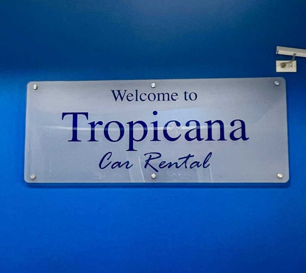 A frosted white sign that reads "Welcome to Tropicana Car Rental" in blue is hung on a blue wall inside the car rental office. Renting a car is one way to get around in St. Maarten.