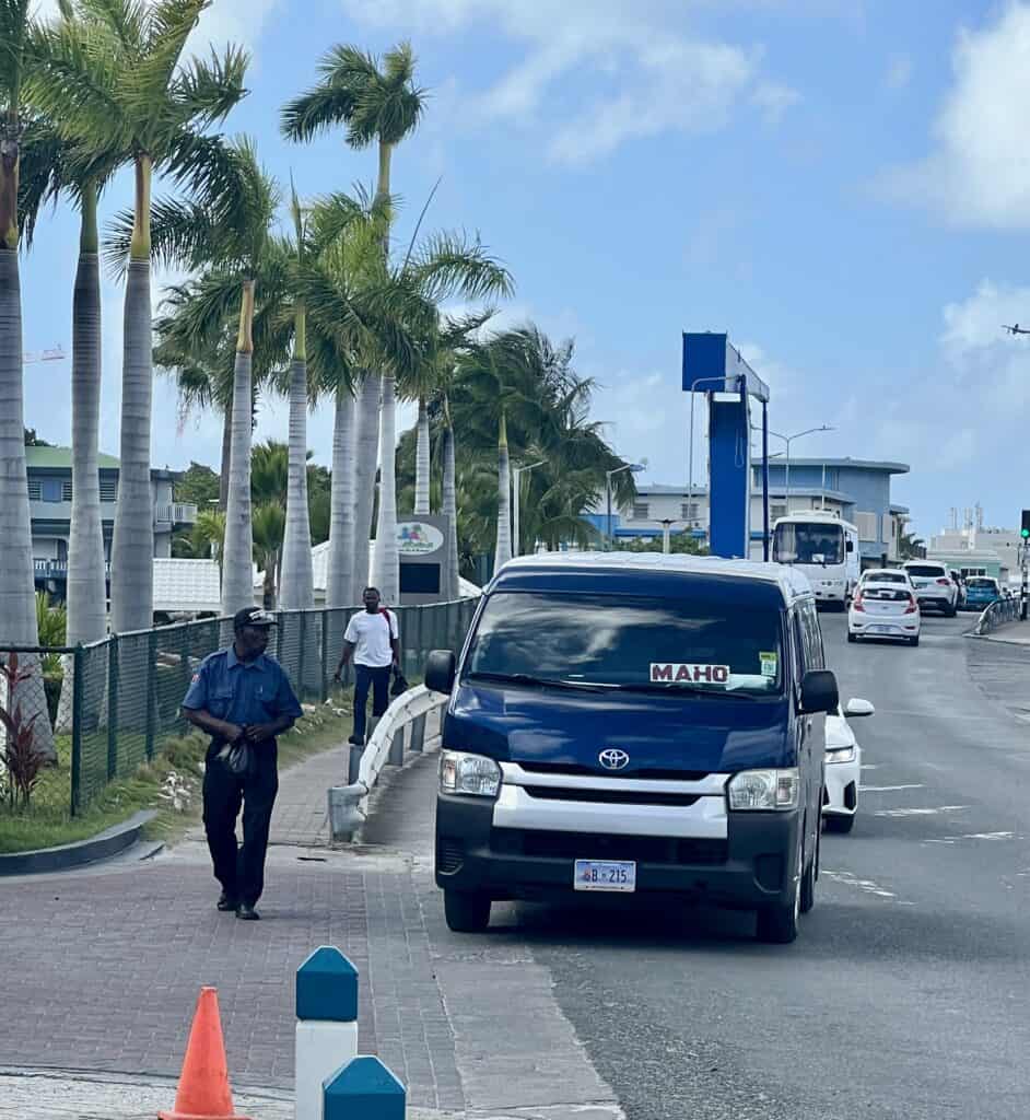 A dark Toyota Van is a bus in St. Maarten. It is driving down the street with a big white sign and the word "MAHO" in red letters. The bus is pulling off the road to pick up a passenger wearing a security uniform.