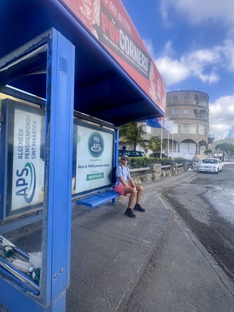 Scott is sitting at a bus stop waiting for the bus in St. Maarten. He is on a bright blue bench with advertisements on the wall behind him and a blue roof over his head. He is wearing a blue shirt, red shorts, a backpack and is smiling at the camera.