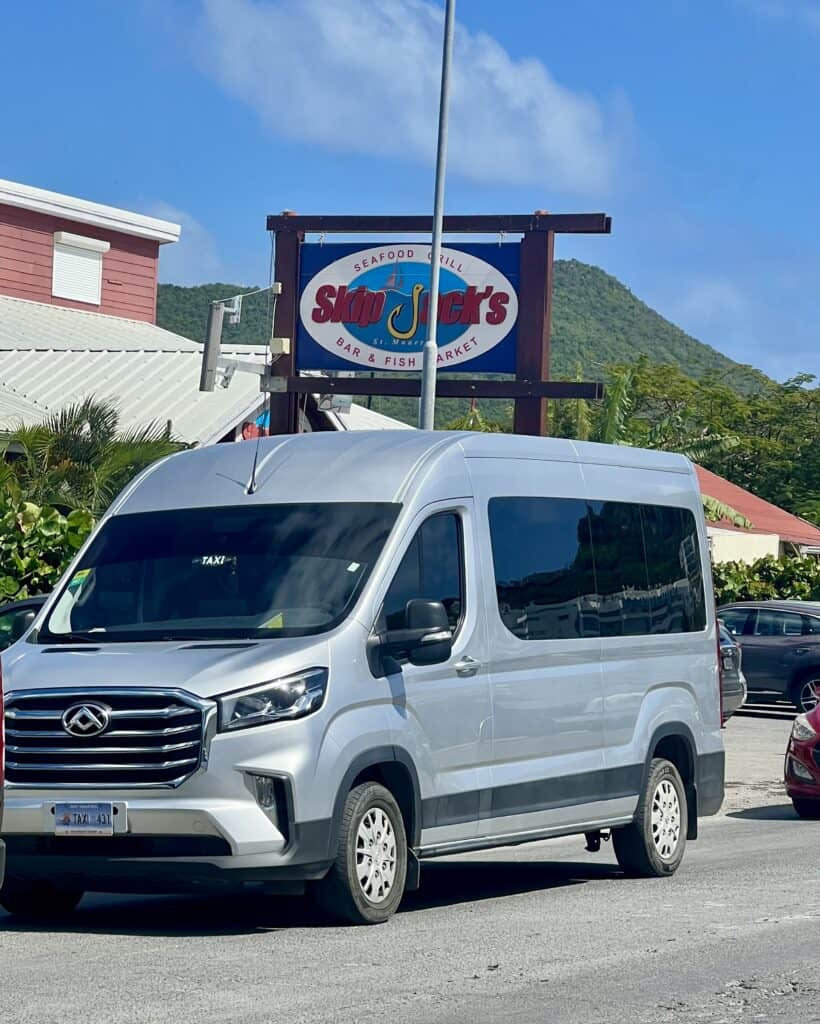 A silver van is driving down the road in St. Maarten. You can tell it is a taxi with its "TAXI" sign at the top of the windshield and the license plate starts with "TAXI". He is stopped in traffic in front of Skip Jacks Bar and Fish Market.
