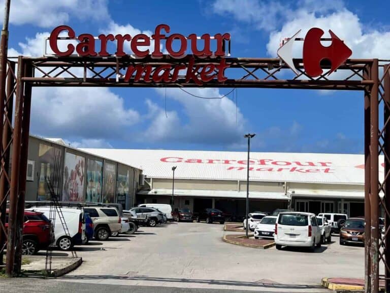 Entrance to the "Carrefour market" grocery store in Cole Bay St. Maarten with cars parked in front beneath a bright blue sky and white clouds. The large red Carrefour signs mark one of the grocery stores serving shoppers on the Dutch side of St. Maarten.