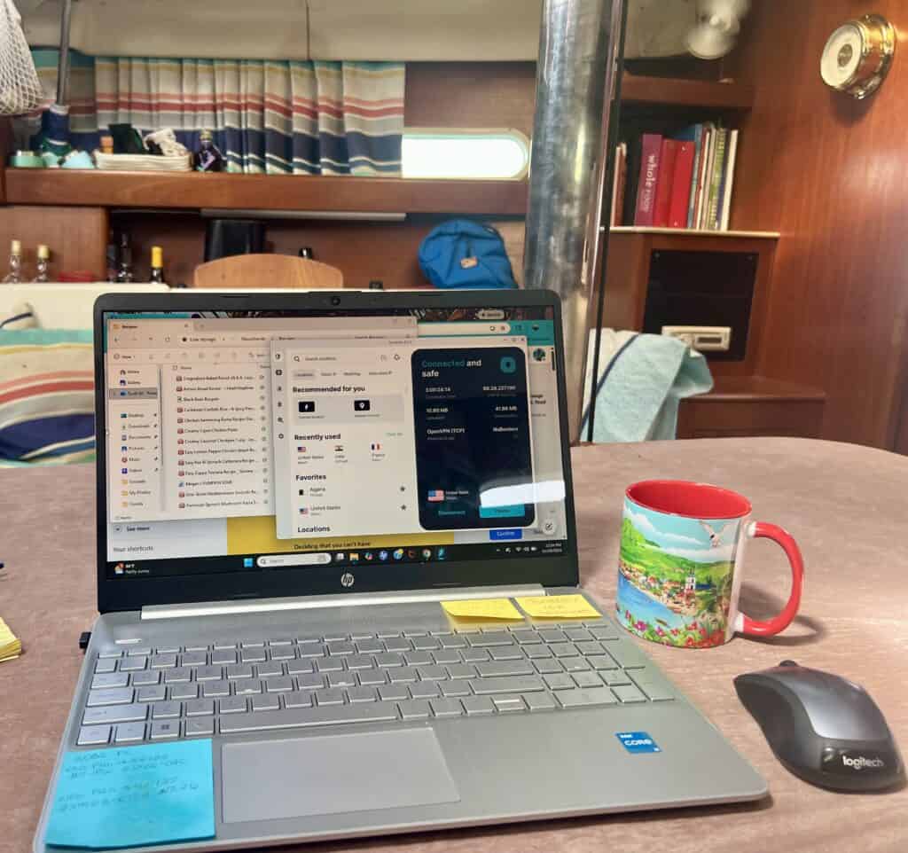 Laptop open inside the cabin of our sailboat Pelagia with Surfshark VPN connected on the screen beside a colorful coffee mug and wireless mouse. Surfshark Review testing setup while working aboard the boat.