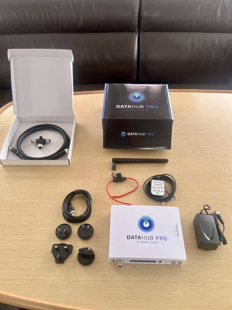 PredictWind DataHub Pro installation kit laid out on a table with the DataHub Pro unit, GPS and GLONASS antenna, external antenna, power supply, cables, adapters, connectors, and packaging. The components shown are included for installing DataHub Pro on a boat.